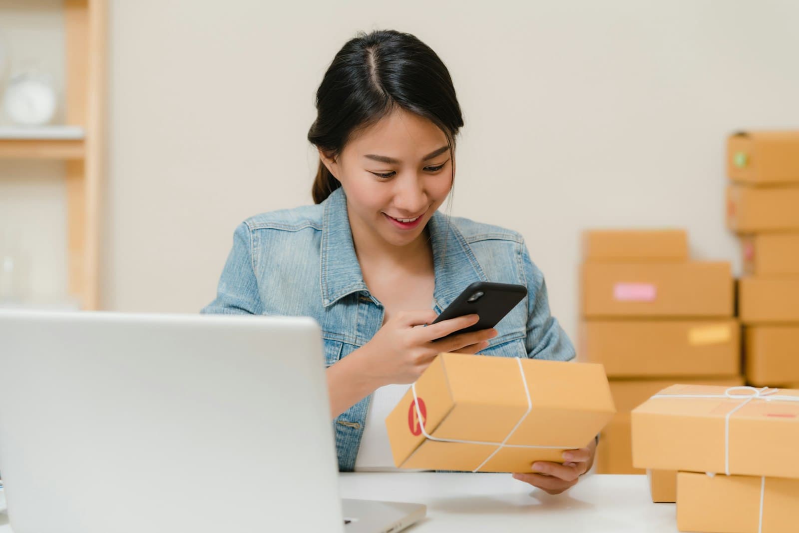 Small-business owner checking an order on her phone beside packed parcels and a laptop
