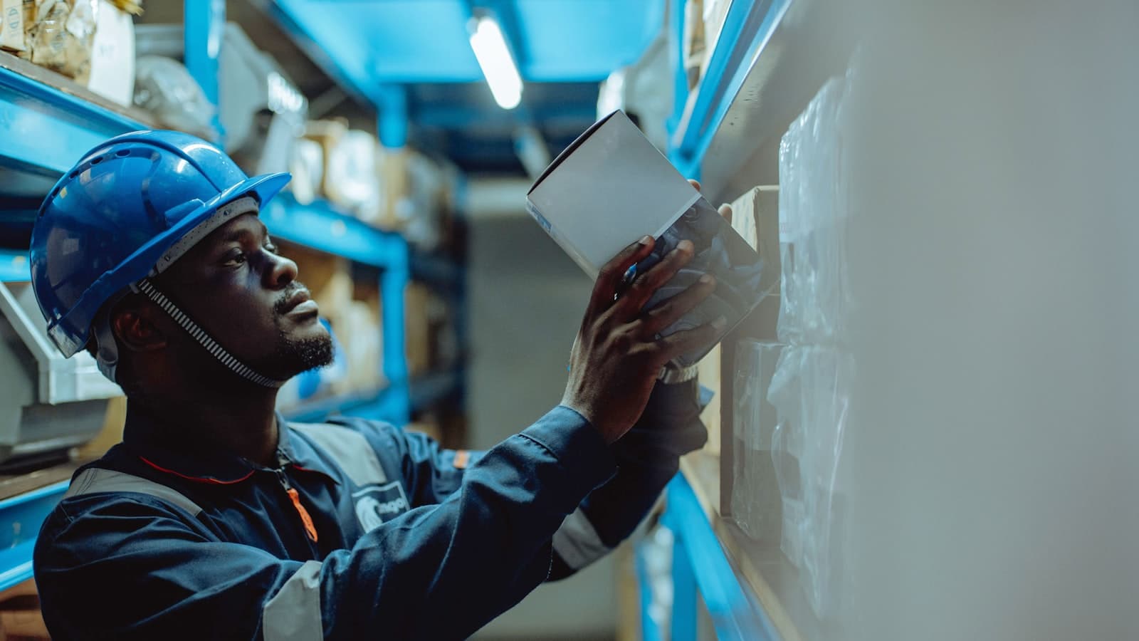 Warehouse worker in a hard hat checking paperwork against stocked shelving